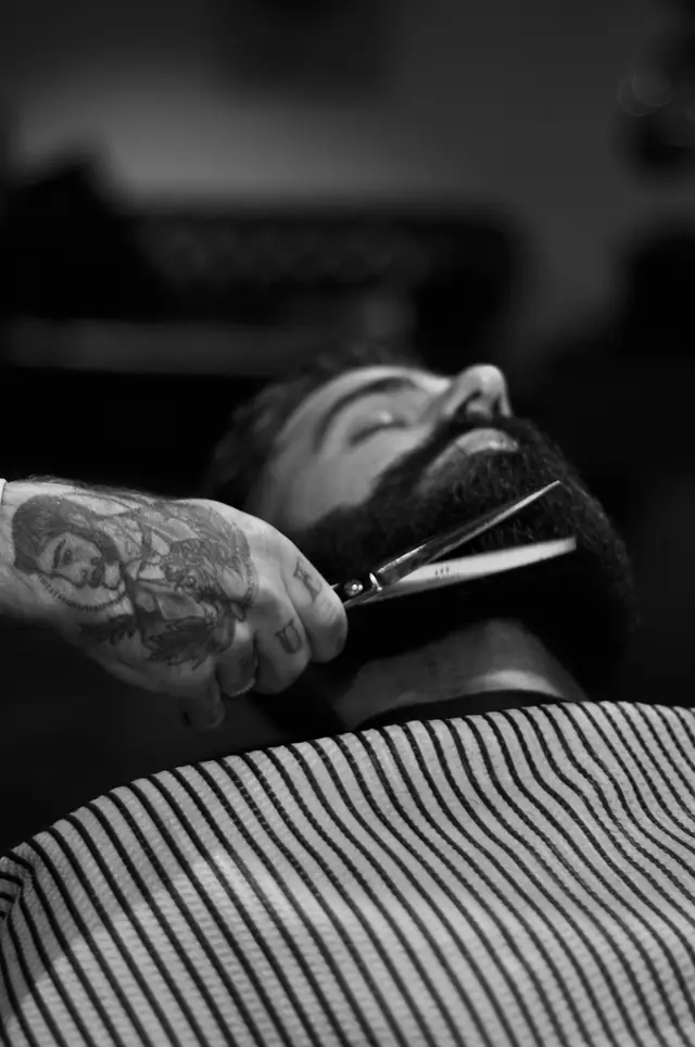 Black and white photo of a barber trimming a customer’s full beard with scissors while he reclines under a striped cape