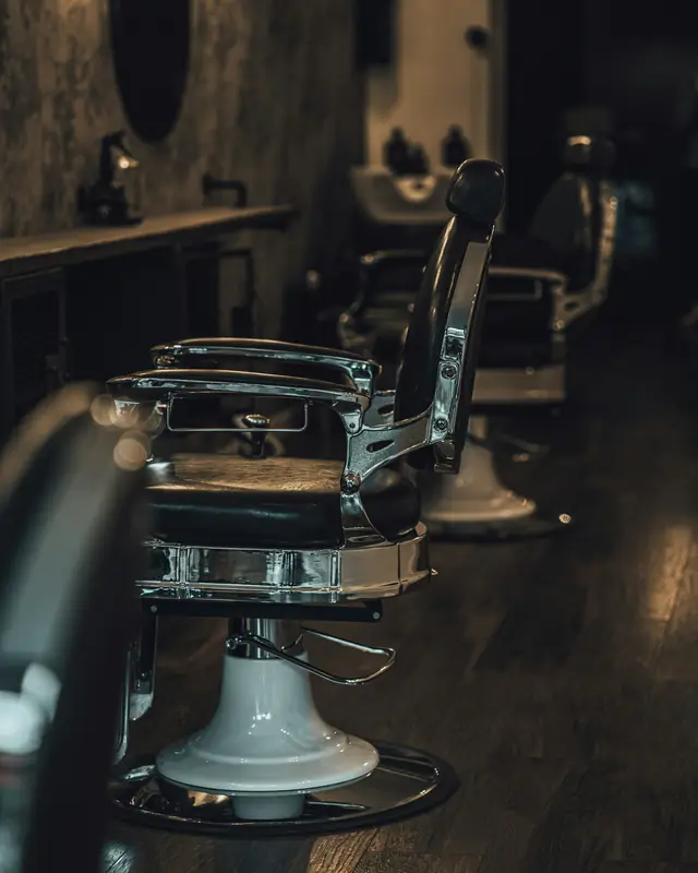 Chrome and black leather barber chairs in a row on a dark wood floor, with a mirror and a sink behind them
