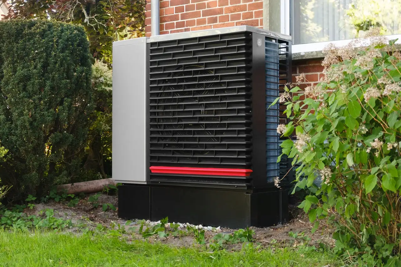 A modern heat pump outdoor unit beside a brick house, surrounded by shrubs and lawn