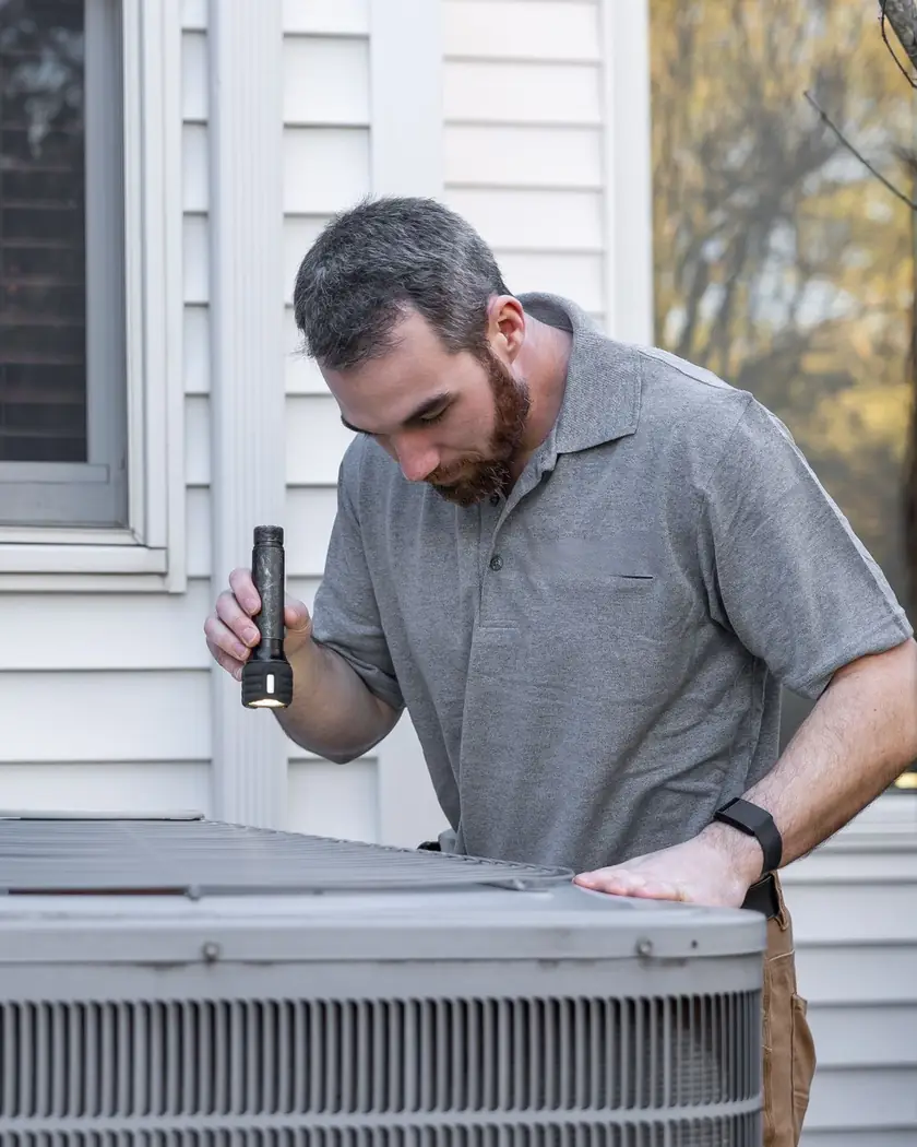 A technician with a flashlight inspecting an outdoor air conditioning unit beside a house