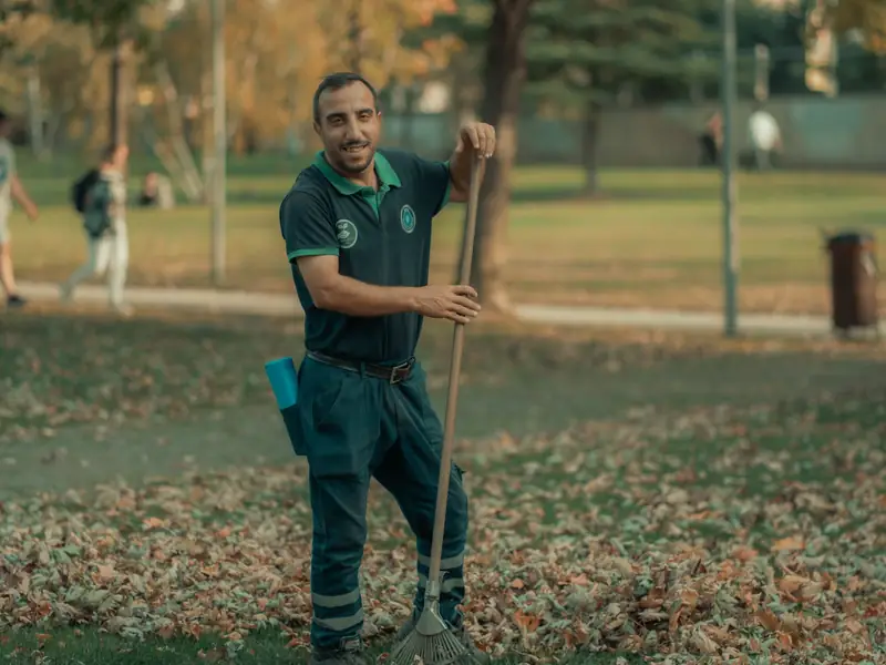 A smiling groundskeeper in a green uniform raking autumn leaves on a lawn