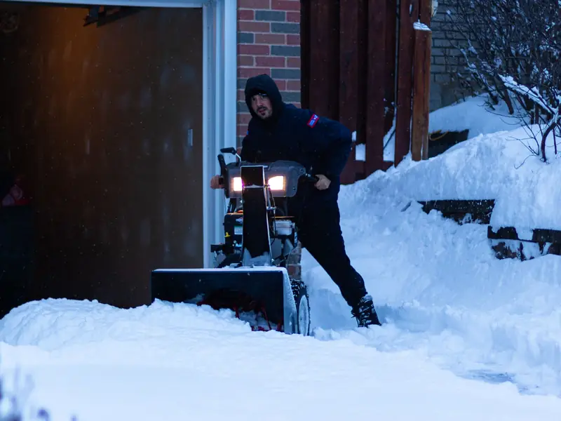 A person clearing a snowy driveway with a snow blower beside a brick house