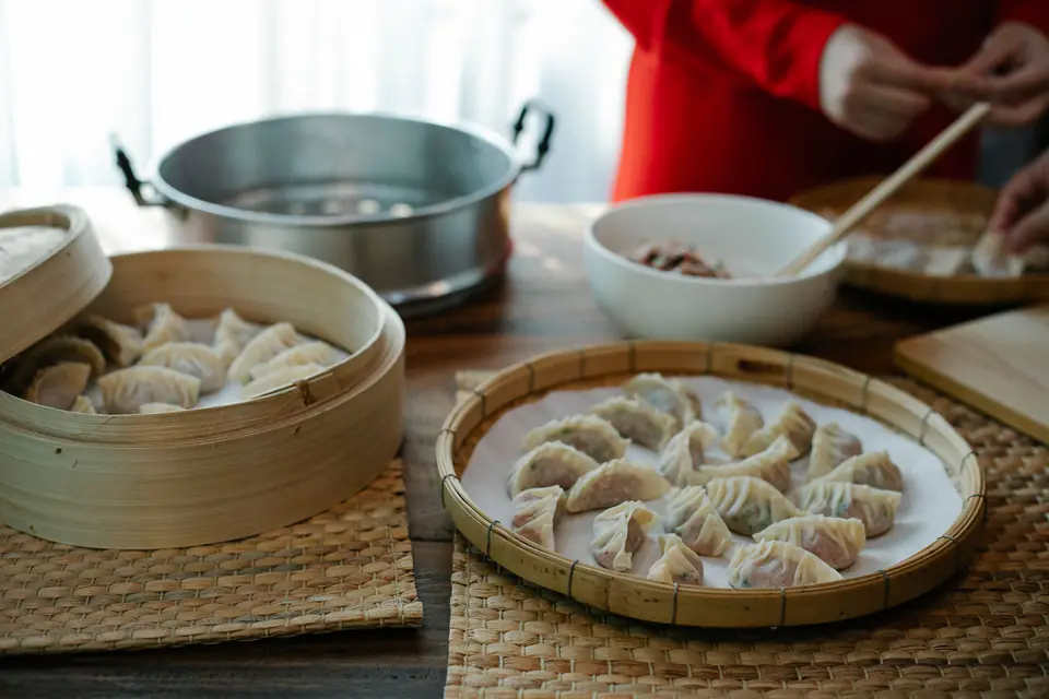 A wooden table with bamboo trays of folded dumplings, a bowl of filling and a steamer
