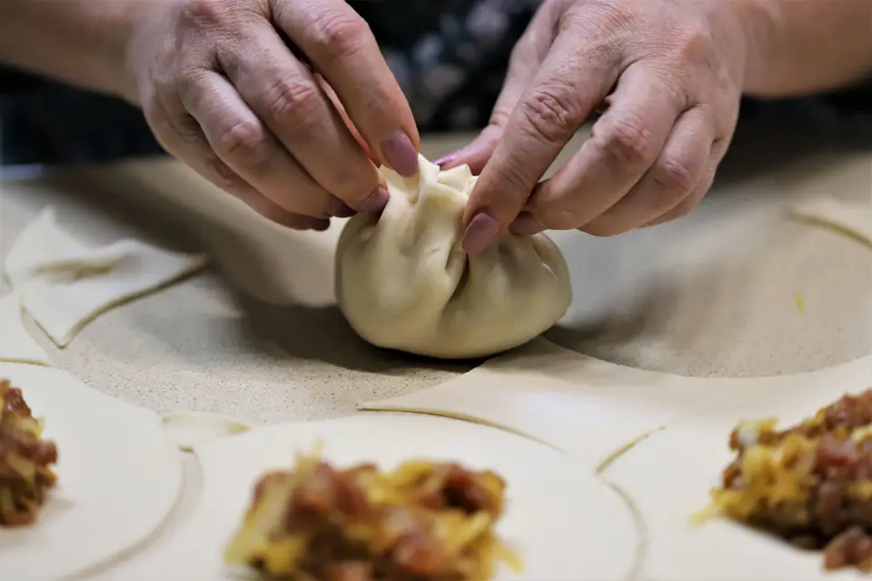 Two hands pinching the pleats of a dumpling closed, with more wrappers and filling on the table