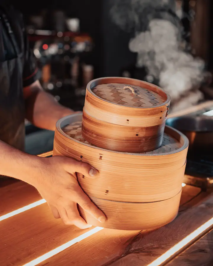 A stack of bamboo steamers with steam rising from the top, carried with both hands