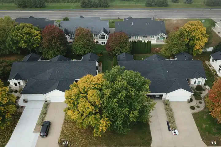 Aerial view of a suburban street with dark asphalt shingle roofs, driveways and autumn trees.